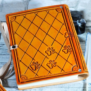 May include: A tan leather-bound journal with a diamond pattern and paw print designs. The journal has a white ribbon bookmark and a metal clasp.