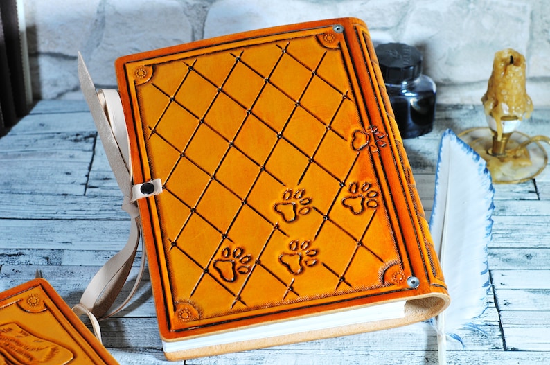 May include: A tan leather-bound journal with a diamond pattern and paw print designs. The journal has a white ribbon bookmark and a metal clasp.