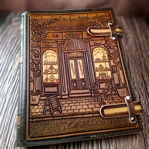 May include: A brown leather journal with a tooled design of a storefront with the text "Patisserie Alexander Paul". The journal has two leather straps with metal buckles.