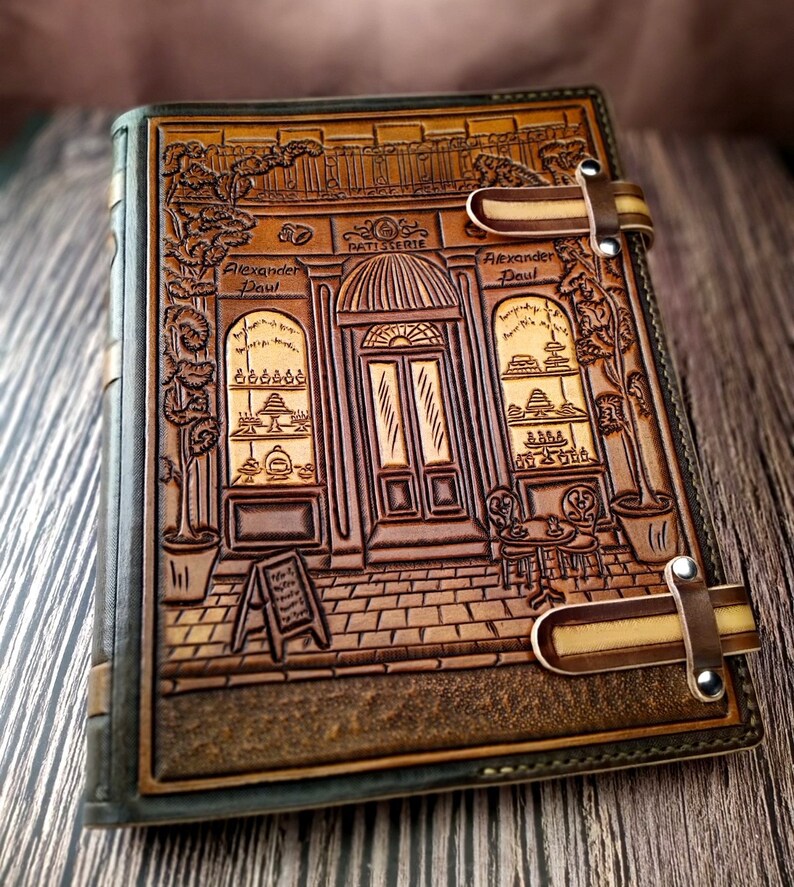 May include: A brown leather journal with a tooled design of a storefront with the text "Patisserie Alexander Paul". The journal has two leather straps with metal buckles.