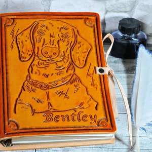 May include: A brown leather journal with a tooled image of a dachshund dog and the name "Bentley" embossed on the front cover. The journal has a ribbon closure and a ring binder.