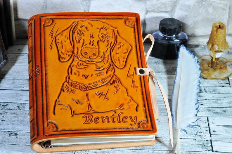 May include: A brown leather journal with a tooled image of a dachshund dog and the name "Bentley" embossed on the front cover. The journal has a ribbon closure and a ring binder.