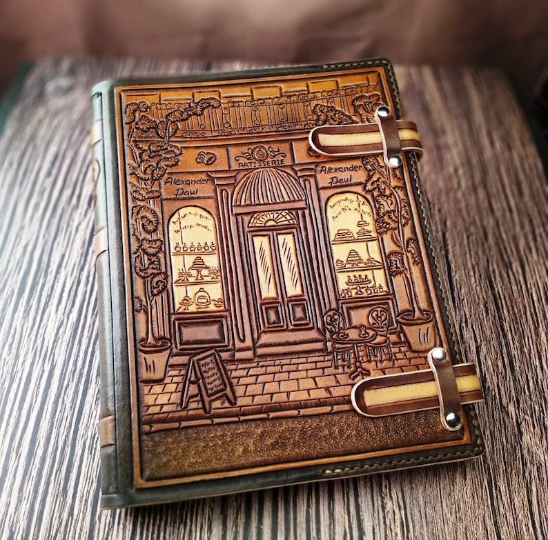 May include: A brown leather journal with a detailed embossed design of a storefront with the text "Patisserie Alexander Paul". The journal has two leather straps with metal clasps.