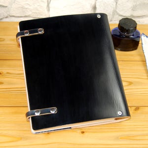 May include: A black leather-bound journal with silver metal clasps and a smooth, polished finish. The journal is lying on a wooden surface.