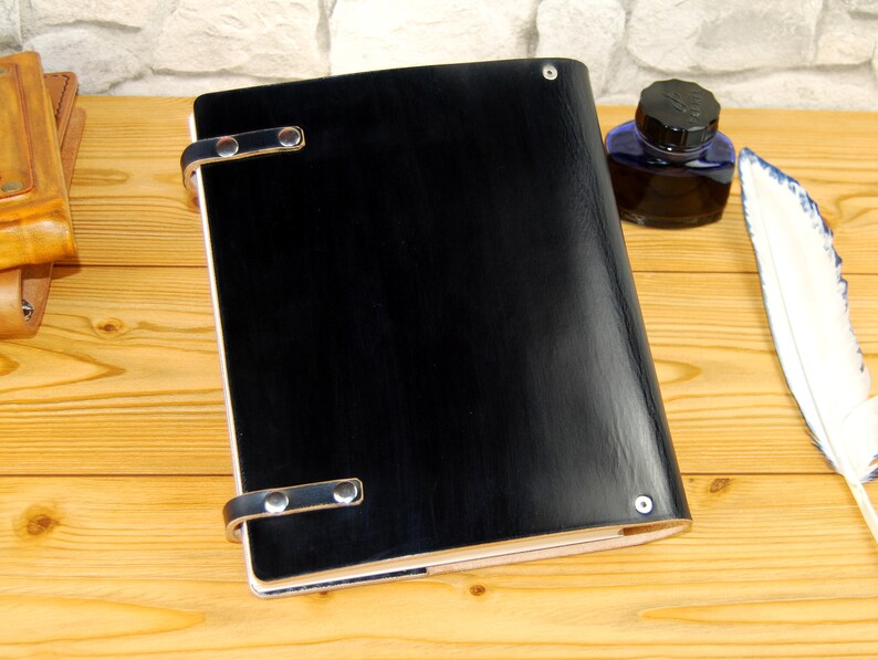 May include: A black leather-bound journal with silver metal clasps and a smooth, polished finish. The journal is lying on a wooden surface.