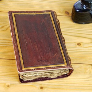 May include: A brown leather-bound journal with gold accents and a worn, vintage look. The journal is closed and lying on a wooden surface.