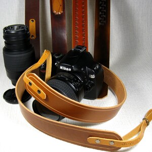May include: A brown leather camera strap with a Nikon camera attached. The strap has a white stitch detail and a metal buckle. The camera is black with a silver lens.