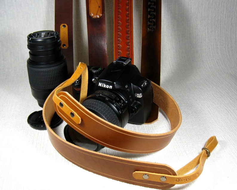 May include: A brown leather camera strap with a Nikon camera attached. The strap has a white stitch detail and a metal buckle. The camera is black with a silver lens.