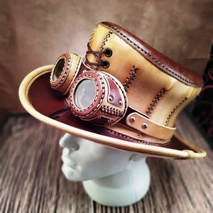May include: A tan and brown leather top hat with a steampunk-style design. The hat has two round goggles attached to the front and a brown leather band with silver rivets.