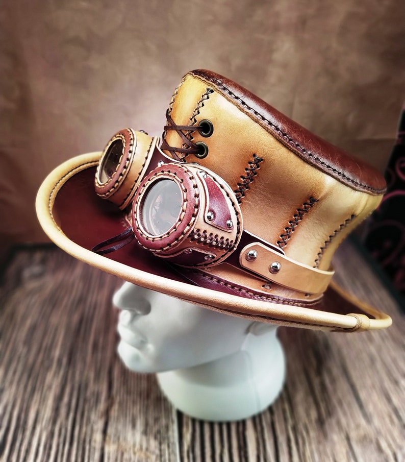 May include: A tan and brown leather top hat with a steampunk-style design. The hat has two round goggles attached to the front and a brown leather band with silver rivets.