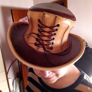 May include: A brown and tan leather top hat with a lace-up detail. The hat is made of two different shades of leather, with a brown brim and a tan crown. The crown has a lace-up detail that is tied with a brown cord.