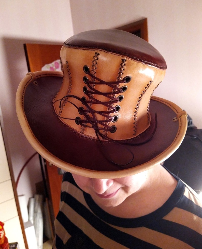 May include: A brown and tan leather top hat with a lace-up detail. The hat is made of two different shades of leather, with a brown brim and a tan crown. The crown has a lace-up detail that is tied with a brown cord.
