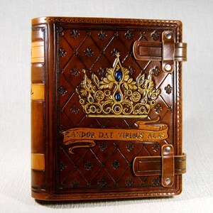 Op de afbeelding: Een bruin lederen gebonden dagboek met een gouden kroon die op de voorzijde is gegraveerd. De kroon is versierd met blauwe edelstenen en de tekst "CANDOR DAT VIRIBUS ALAS" staat op een banner eronder.