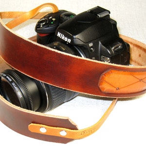 May include: A brown leather camera strap with a tan leather accent. The strap is wrapped around a black camera with a lens.
