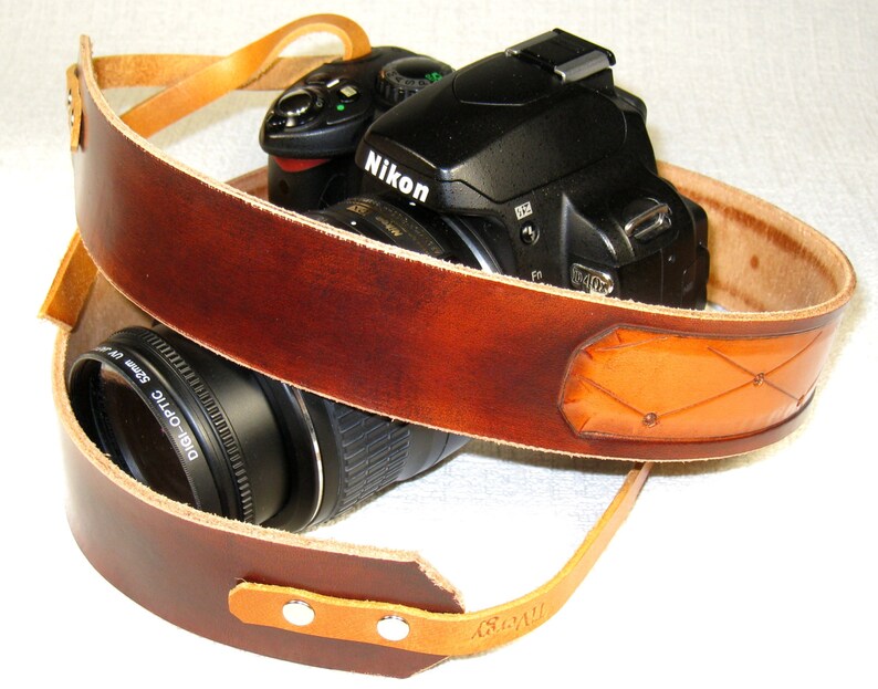 May include: A brown leather camera strap with a tan leather accent. The strap is wrapped around a black camera with a lens.