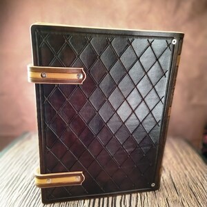 May include: A brown leather journal with a diamond pattern embossed on the cover. The journal has two brass clasps with a cream colored leather trim. The journal is sitting on a wooden surface.