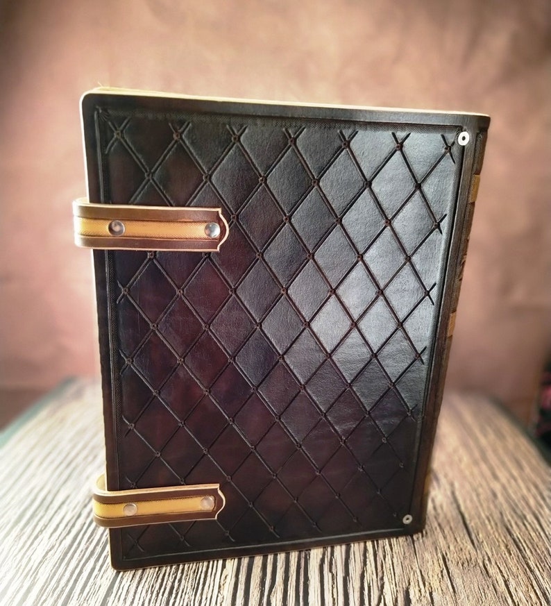 May include: A brown leather journal with a diamond pattern embossed on the cover. The journal has two brass clasps with a cream colored leather trim. The journal is sitting on a wooden surface.