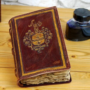May include: A brown leather-bound journal with gold accents and a decorative crest on the front cover. The journal is open to reveal blank pages. A white feather quill pen and a bottle of ink are also in the image.