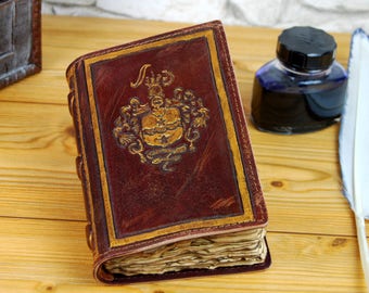 Pode incluir: Um diário encadernado em couro marrom com detalhes dourados e um brasão decorativo na capa. O diário está aberto para revelar páginas em branco. Também há uma pena de pena branca e um frasco de tinta na imagem.