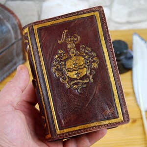 May include: A brown leather-bound journal with gold accents and a gold embossed design on the front cover. The design features a crest with a crown and other decorative elements.