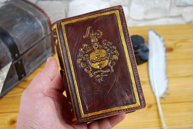 May include: A brown leather-bound journal with gold accents and a gold embossed design on the front cover. The design features a crest with a crown and other decorative elements.