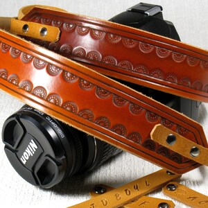 May include: A brown leather camera strap with a decorative embossed pattern. The strap is adjustable and has a metal buckle.