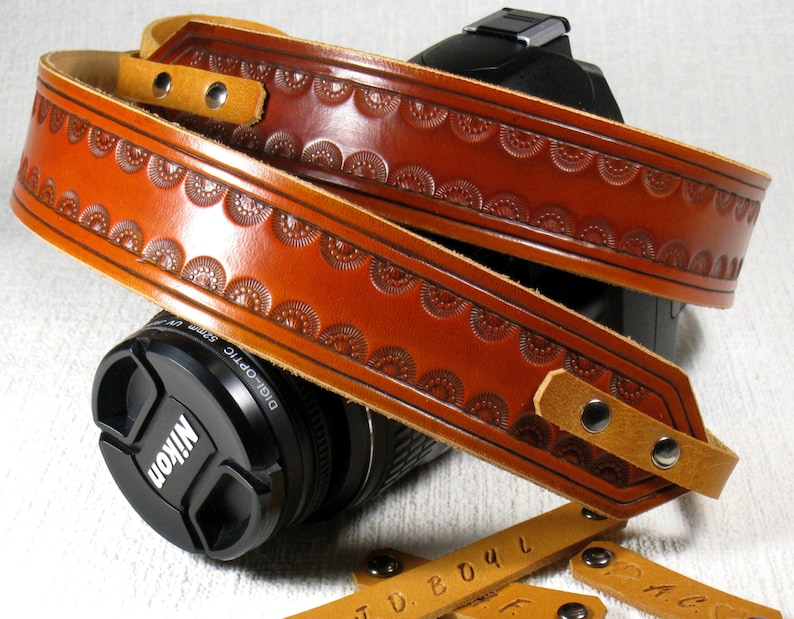 May include: A brown leather camera strap with a decorative embossed pattern. The strap is adjustable and has a metal buckle.