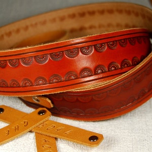 May include: A brown leather guitar strap with a decorative embossed pattern. The strap is folded in half and has two brown leather tags with the text "3H" and "KOAL" on them.