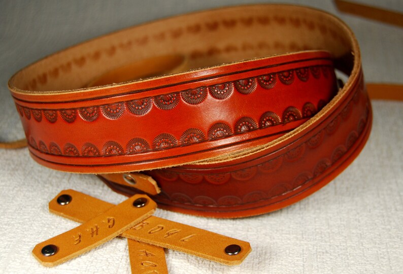 May include: A brown leather guitar strap with a decorative embossed pattern. The strap is folded in half and has two brown leather tags with the text "3H" and "KOAL" on them.