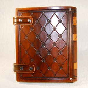 Op de afbeelding: Een bruin lederen gebonden dagboek met een decoratief reli&euml;fpatroon van fleur-de-lis en een sluiting met een band. Het dagboek heeft een rug met een titel en een decoratief ontwerp.