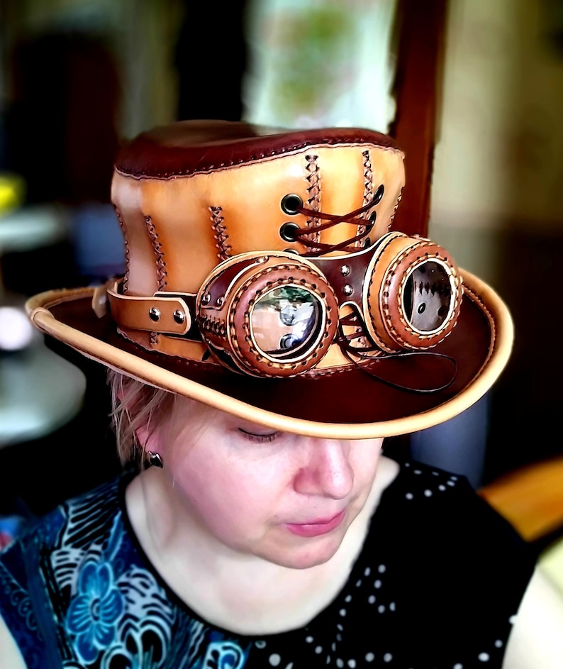 May include: A brown leather top hat with a tan leather band and goggles. The hat has a lace-up detail on the side and the goggles have round lenses.