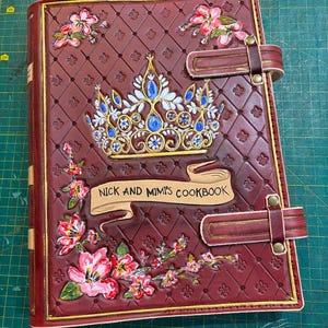 Op de afbeelding: Een bordeauxrood lederen kookboek met een gouden kroonontwerp, bloemmotieven en de tekst "Nick and Mimi's Cookbook" op een banner.
