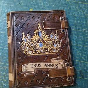 Op de afbeelding: Een bruin lederen gebonden dagboek met een gouden kroon en blauwe juwelen, een banner met de tekst "Unus Annus" en twee leren banden.