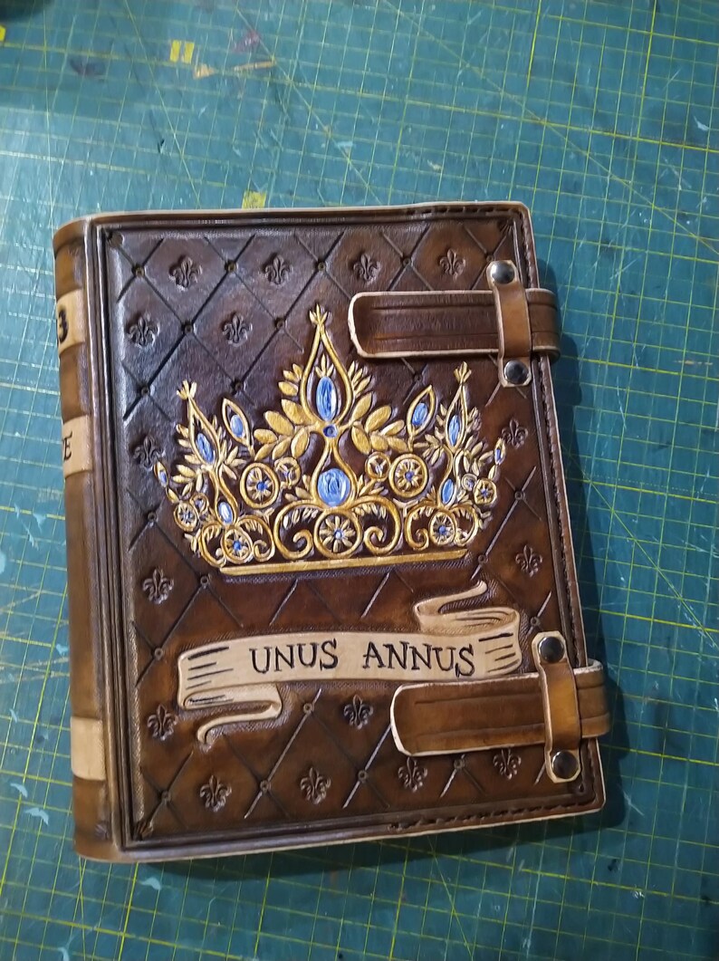 Op de afbeelding: Een bruin lederen gebonden dagboek met een gouden kroon en blauwe juwelen, een banner met de tekst "Unus Annus" en twee leren banden.
