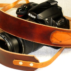 May include: A brown leather camera strap with a tan leather patch and a Nikon camera attached. The strap has a woven design and the camera has a lens attached.