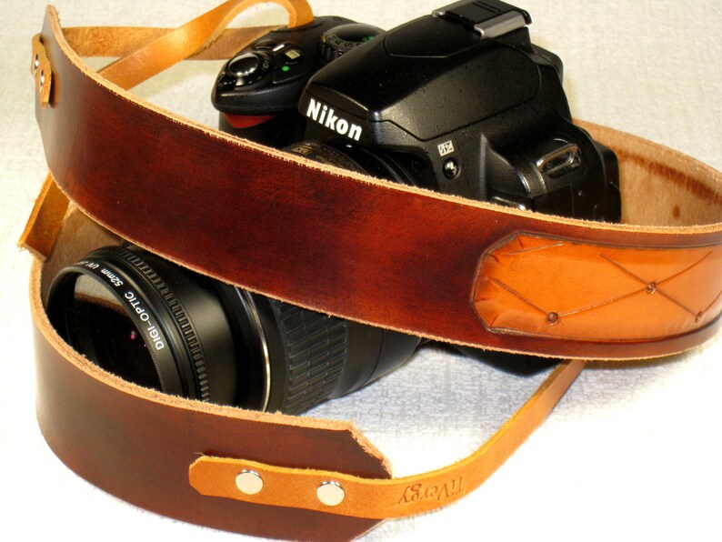 May include: A brown leather camera strap with a tan leather patch and a Nikon camera attached. The strap has a woven design and the camera has a lens attached.