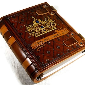 Op de afbeelding: Een bruin lederen gebonden dagboek met een gouden kroon en blauwe juwelen op de omslag. Het dagboek heeft een decoratief reli&euml;fpatroon en de tekst "Candor Dat Virtus Alas".