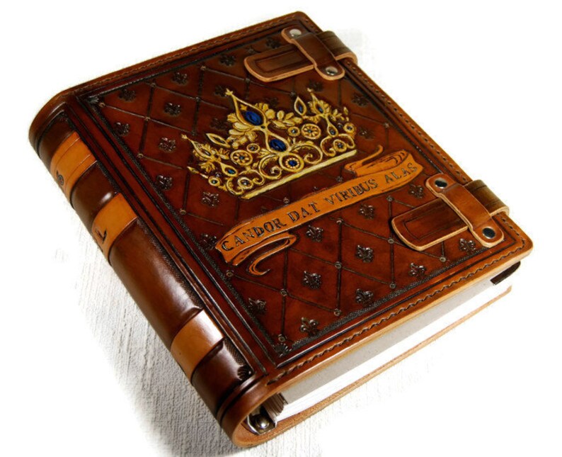 Op de afbeelding: Een bruin lederen gebonden dagboek met een gouden kroon en blauwe juwelen op de omslag. Het dagboek heeft een decoratief reli&euml;fpatroon en de tekst "Candor Dat Virtus Alas".