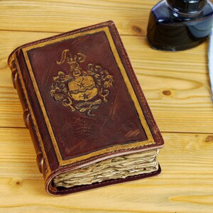 May include: A brown leather-bound journal with gold accents and a decorative crest on the front cover. The journal is closed and sitting on a wooden surface.