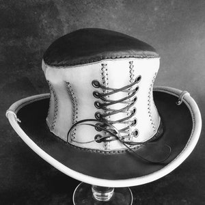 May include: A white and brown leather hat with a corset-style lacing detail. The hat is sitting on a clear glass stand.