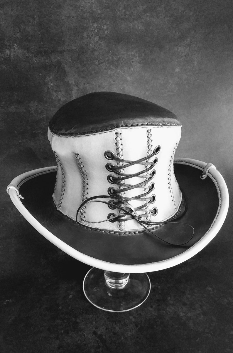 May include: A white and brown leather hat with a corset-style lacing detail. The hat is sitting on a clear glass stand.