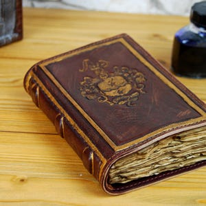 May include: A brown leather-bound journal with gold accents and an embossed design. The journal is closed and sitting on a wooden surface.