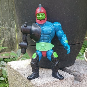May include: A green and blue action figure with a red helmet and a skull and crossbones on its belt. The figure is holding a black weapon in its right hand.