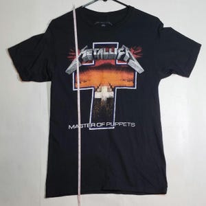 Puede incluir: Camiseta negra con el logo de Metallica en plata y rojo, y las palabras "MASTER OF PUPPETS" debajo de un gráfico en forma de cruz. La camiseta está colgada en una percha negra.