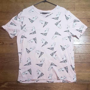 Può includere: Una maglietta rosa chiaro con un motivo ripetuto di Snoopy del fumetto Peanuts. Snoopy è raffigurato con uno striscione che dice "PEANUTS" in nero. La maglietta ha un girocollo e maniche corte.