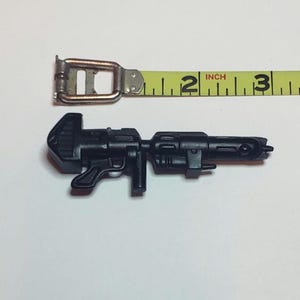 May include: A black plastic toy gun, approximately 3 inches long, is displayed next to a yellow measuring tape. The gun has a detailed design with various protrusions and a handle. The measuring tape shows inch measurements.