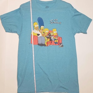 May include: A light blue t-shirt featuring the Simpsons family on a red couch. The graphic includes Homer, Marge, Bart, Lisa, and Maggie, along with the show's logo. The shirt is made of a soft material.