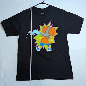 May include: Black t-shirt featuring a cartoon character with large, orange and gray eyes, wearing an orange shirt and blue shorts. The character is depicted with water spraying from its face, set against a green and purple burst.