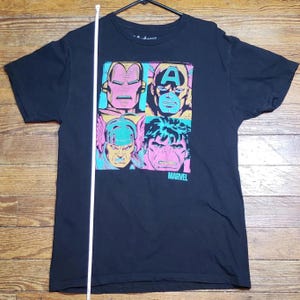 May include: Black t-shirt featuring a colorful graphic of four Marvel superheroes: Iron Man, Captain America, Thor, and Hulk. The Marvel logo is printed below the images. The shirt is hanging on a black hanger.