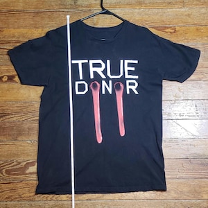 Puede incluir: Camiseta negra con el texto "TRUE DONOR" en letras blancas. Debajo del texto hay dos diseños verticales rosas estilizados de gotas de sangre. La camiseta está colgada de una percha negra.